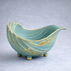 Op de afbeelding: Een turquoise decoratieve kom met een golvend ontwerp en gouden accenten. De kom heeft drie kleine voetjes en gouden bladpatronen. De rand heeft een geschulpte rand. Een uniek stuk voor de interieurdecoratie.