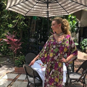 May include: A woman wearing a long, flowing, multi-coloured patterned dress with a V-neckline. The dress has a geometric pattern with shades of purple, green, and brown. She is standing on a patio with a table and chairs in the background.