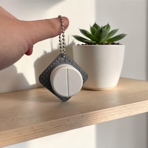 Puede incluir: Un llavero gris y blanco con forma de pastilla con una cadena de bolas. El llavero tiene un borde cuadrado texturizado y un centro blanco en forma de pastilla con una línea vertical. Una pequeña planta suculenta en una maceta blanca está en una estantería de madera.