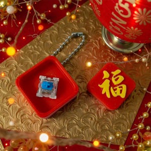 Könnte beinhalten: Ein roter, quadratischer Schlüsselanhänger mit einer silbernen Kette, der einen transparenten und blauen mechanischen Tastaturschalter hält. Daneben befindet sich ein rotes Quadrat mit gelben chinesischen Schriftzeichen. Der Hintergrund ist gold und rot mit dekorativen Lichtern.