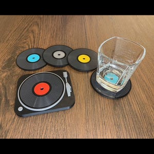 Op de afbeelding: Een set van vinylplaten-vormige onderzetters en een onderzetter in de vorm van een platenspeler. De onderzetters zijn zwart met gekleurde middellabels in blauw, geel en rood. Een helder vierkant glas staat op een onderzetter.