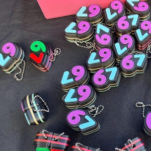 Puede incluir: Una colección de llaveros coloridos con los números 67 y 29. Los números son de varios colores, incluyendo morado, azul y verde, y están unidos a pequeñas cadenas de metal.