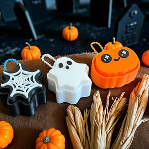 Pode incluir: Três pequenos recipientes com tampa em forma de ícones de Halloween: uma caixa preta com tampa de teia de aranha branca, uma caixa branca em forma de fantasma e uma caixa laranja em forma de abóbora com um rosto sorridente e alças.