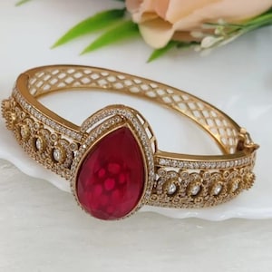 Op de afbeelding: Goudkleurige armband met een grote, druppelvormige, robijnrode edelsteen. De armband heeft ingewikkelde filigraan en kleine, sprankelende accenten. Het ontwerp omvat een rasterpatroon op de band, waardoor een elegant en sierlijk sieraad ontstaat.