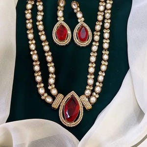 May include: A gold-toned necklace and earring set featuring red teardrop-shaped gemstones. The necklace has a double-strand design with white, oval-shaped accents. The earrings mirror the necklace's gemstone design. The jewelry is displayed on a dark green background with white fabric.