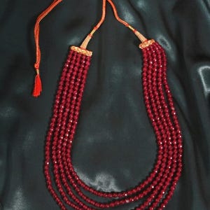 May include: A multi-strand necklace featuring small, faceted red beads. The necklace has an adjustable orange cord with a tassel and gold-colored accents. The beads are arranged in multiple strands, creating a layered effect. The background is a dark, textured fabric.