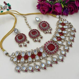 May include: A gold-toned jewelry set featuring a necklace, earrings, and a headpiece. The necklace and earrings have a combination of red and clear stones. The headpiece has a single red stone and clear stones. The jewelry is set against a white background.