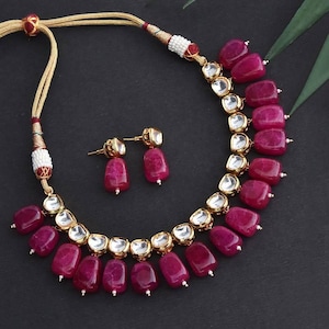 Puede incluir: Un conjunto de collar y pendientes con piedras rectangulares grandes de color magenta. El collar tiene detalles dorados y un par de pendientes a juego. Las joyas están sobre un fondo oscuro.
