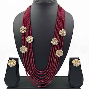 Puede incluir: Un collar de cuentas burdeos de varias hebras con pendientes a juego. El collar presenta detalles florales dorados. Las joyas se exhiben sobre una cabeza de maniquí negra y soportes para pendientes sobre un fondo blanco.