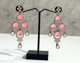 Può includere: Un paio di orecchini a lampadario con parte superiore nera, pietre centrali rosa e dettagli trasparenti a forma di goccia. Gli orecchini sono esposti su un supporto nero su sfondo neutro. Estetica vintage o antica.