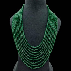 Puede incluir: Un collar de múltiples hilos compuesto por pequeñas cuentas redondas de color verde esmeralda. El collar se exhibe sobre un busto de maniquí negro sobre un fondo negro. Las cuentas caen en cascada en un diseño en capas, creando un aspecto completo y elegante.