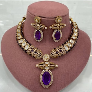 Op de afbeelding: Een goudkleurige ketting- en oorbellenset met een paarse ovale edelsteen hanger. De ketting en oorbellen hebben ingewikkelde details met heldere en paarse stenen. De sieraden worden getoond op een roze mannequinhoofd.