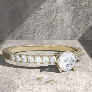 May include: A gold ring with a large, round, clear diamond center stone. The band is also set with smaller, clear diamonds. The ring is on a light gray surface.