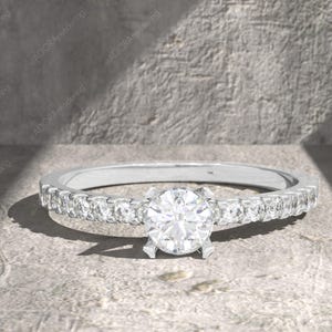 May include: A silver engagement ring with a large, round, clear gemstone center and smaller gemstones set along the band. The ring is set on a textured, light gray surface.