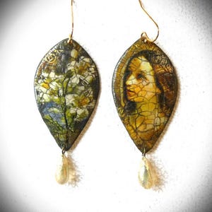 May include: Pair of leaf-shaped earrings with gold-coloured ear wires. One earring features a floral design with white and yellow flowers, the other a portrait of a woman. Each earring has a pearlescent teardrop charm.