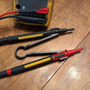 May include: A yellow and black Fluke multimeter with red and black test leads. Two black test probes with yellow labels and a red probe tip are also visible. The items are on a wooden surface.