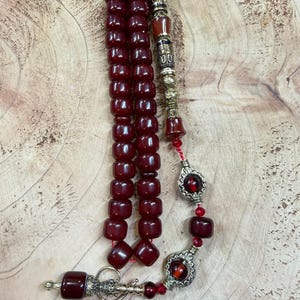 May include: A maroon prayer bead necklace with a decorative silver-toned metal divider and accents. The beads are a deep red color and are strung on a red cord. The necklace has a decorative tassel and a silver-toned metal pendant.