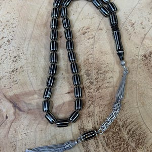 Peut inclure: Un chapelet noir et blanc avec un pompon argenté. Les perles oblongues sont rayées, reliées par une chaîne argentée. Le pompon est fait de fils d'argent et possède un capuchon argenté décoratif.