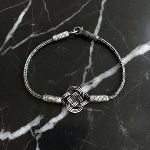 May include: A silver bracelet with a Celtic knot design. The bracelet features a woven chain and decorative end pieces. The knot is the focal point, and the bracelet is set against a black marble surface.