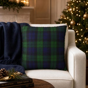 May include: A square throw pillow featuring a dark green and navy blue plaid tartan pattern. The fabric displays a classic intersecting grid design with thin black lines separating the larger blocks of color, creating a balanced and uniform textile appearance.