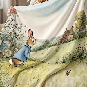 May include: A soft throw blanket featuring a printed illustration of a brown rabbit wearing a blue jacket standing in a grassy field. The scene includes a stone cottage, tall wildflowers, and a small butterfly against a pale sky background.