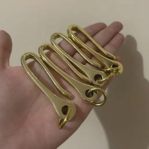 May include: A set of four polished gold-tone metal fishhook key clasps. Each clasp features an elongated, curved hook design with a circular eyelet at the base, and each is equipped with a small, circular metal jump ring attached to the eyelet.