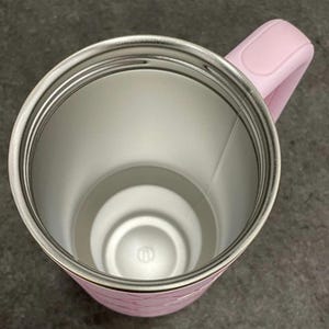 May include: A pink insulated tumbler with a stainless steel interior. The tumbler has a matching pink handle and a decorative pattern on the exterior. The interior is silver and the handle is curved for easy gripping. The tumbler is designed to keep beverages hot or cold.