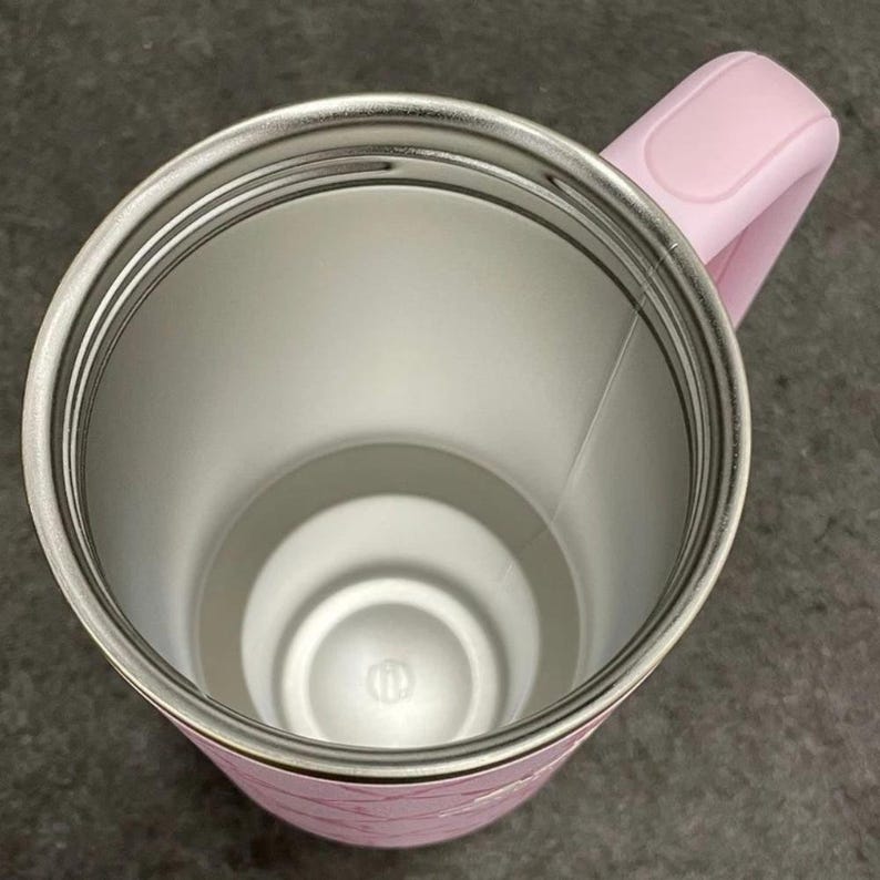 May include: A pink insulated tumbler with a stainless steel interior. The tumbler has a matching pink handle and a decorative pattern on the exterior. The interior is silver and the handle is curved for easy gripping. The tumbler is designed to keep beverages hot or cold.