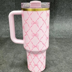 May include: Pink insulated tumbler with a handle, featuring a repeating pattern of pink bows and ribbons. The tumbler has a gold band near the top and a clear lid with a pink slider. The handle and lid are also pink.