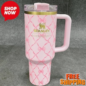 May include: A pink Stanley Loveshackfancy travel mug with a handle. The mug features a bow pattern and the Stanley logo. The mug has a clear lid and a gold band near the top. The mug is designed for cold and hot beverages.
