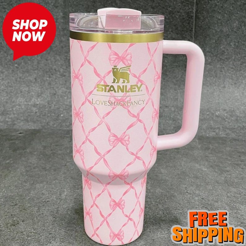 May include: A pink Stanley Loveshackfancy travel mug with a handle. The mug features a bow pattern and the Stanley logo. The mug has a clear lid and a gold band near the top. The mug is designed for cold and hot beverages.