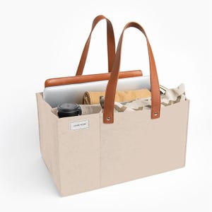 May include: A beige tote bag with brown leather handles and accents. The bag contains a laptop, a rolled-up blanket, and a black tumbler. The text "CHASE HOME" is visible on the front of the bag.