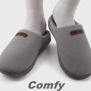 Könnte beinhalten: Graue Hausschuhe in Waffelstrick-Optik mit einem braunen Etikett mit der Aufschrift "ofoot". Die Hausschuhe werden mit weißen Socken getragen. Das Wort "Comfy" ist unten in Grau aufgedruckt.