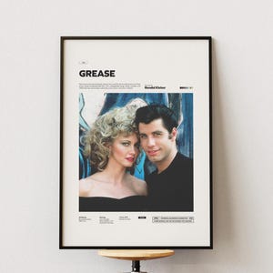 Può includere: Poster incorniciato del film "Grease". La cornice è nera e il poster mostra una foto dei due attori principali. Il titolo "Grease" è in alto, con testo sotto l'immagine. Lo sfondo è bianco.
