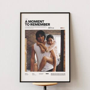 May include: A framed movie poster for "A Moment to Remember" featuring a man carrying a woman. The poster has a black frame and is mounted on a white wall. The title is in bold, black text at the top.
