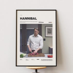 May include: Framed poster for the TV show "Hannibal." The poster shows a man in a white shirt cutting meat on a wooden board. The poster includes the show's title, credits, and a rating.