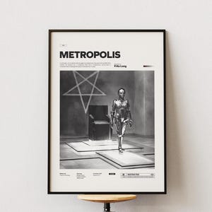 May include: Black and white framed poster featuring the word "METROPOLIS" at the top. The poster depicts a robot walking towards the viewer, with a pentagram and chair in the background. The poster is mounted on a white wall.