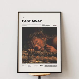 May include: A framed movie poster for "Cast Away" featuring a distressed man lying on a pile of debris. The poster has the movie title in bold white letters at the top, with the director's name and other details below.