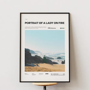 Puede incluir: Póster enmarcado con el título "PORTRAIT OF A LADY ON FIRE". La imagen presenta una escena de playa con una persona con un vestido verde. El cielo es azul claro y la arena es de color arena. El póster está en un marco negro.