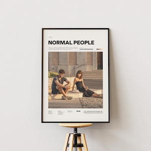 Puede incluir: Un póster enmarcado con el título "NORMAL PEOPLE" en letras negras en negrita. El póster presenta una fotografía de un hombre y una mujer sentados al aire libre. El hombre lleva una camiseta oscura y pantalones cortos, y la mujer un vestido negro.