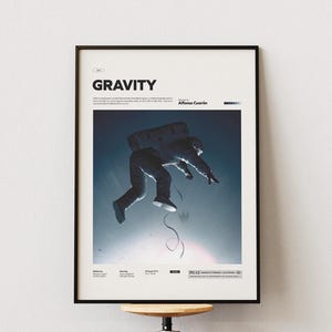 Może przedstawiać: Czarny plakat z napisem "GRAVITY" w białych literach. Obraz przedstawia astronautę w ciemnym skafandrze kosmicznym unoszącego się na tle niebiesko-białego gradientu. Plakat zawiera nazwisko reżysera i inne szczegóły.