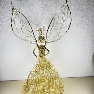 Può includere: Una scultura in filo metallico color oro raffigurante una figura alata con una gonna arrotondata e voluminosa realizzata in filo avvolto. La figura presenta un busto dettagliato, braccia posizionate sui fianchi e grandi ali complesse.