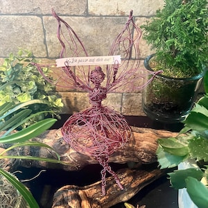Puede incluir: Una escultura de hada de alambre hecha a mano en un tono burdeos intenso. El hada tiene alas extendidas y un delicado vestido de alambre. Un pequeño letrero dice "Ich passe auf dich auf!". El hada se exhibe sobre madera flotante.