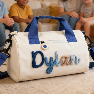 May include: A white duffel bag with blue accents, a blue handle, and a blue shoulder strap. The name "Dylan" is embroidered on the front in blue and tan yarn. The bag has a zipper and black clips.