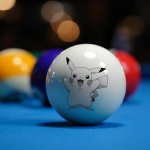 Op de afbeelding: Een witte biljartbal met een Pikachu-afbeelding, geplaatst voor andere gekleurde biljartballen op een blauwe pooltafel. Het Pikachu-ontwerp is grijs en toont het personage met opgeheven armen. De ballen zijn scherp, met een wazige achtergrond.