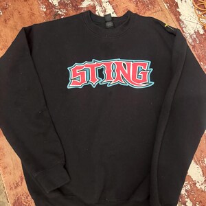Op de afbeelding: Zwarte crewneck sweatshirt met het woord "STING" in een rood en lichtblauw grafisch ontwerp over de borst. De sweatshirt heeft een geribbelde kraag, manchetten en zoom. Het kledingstuk is gemaakt van een zacht materiaal.