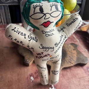 May include: A beige fabric doll with a green ribbon hair accessory, featuring hand-drawn facial features and black text printed across the body listing various physical ailments including Liver Spots, Flabby Arms, Hot Flashes, and Pot Belly.