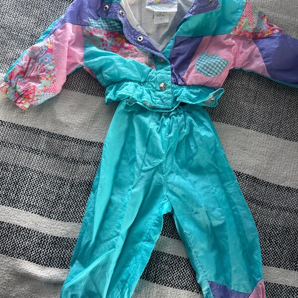 May include: A retro two-piece tracksuit in a bright colour scheme. The jacket has a patchwork design with pink, purple, and teal panels, and a ruffled waist. The matching trousers are a solid teal colour. The tracksuit is made of a shiny, lightweight material.