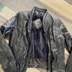 Puede incluir: Chaqueta de cuero negra con cuello alzado y cremallera. La chaqueta tiene un diseño acolchado en los hombros y las mangas. El interior está forrado con un material azul oscuro y difuso. La etiqueta interior dice "Leather Warehouse".