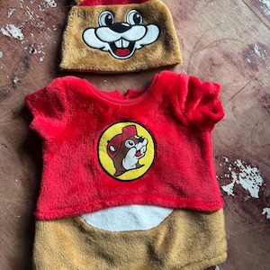 May include: A red and brown baby outfit with a matching hat. The outfit features a beaver logo on the chest and a brown bottom. The hat is brown with a red top and beaver face.